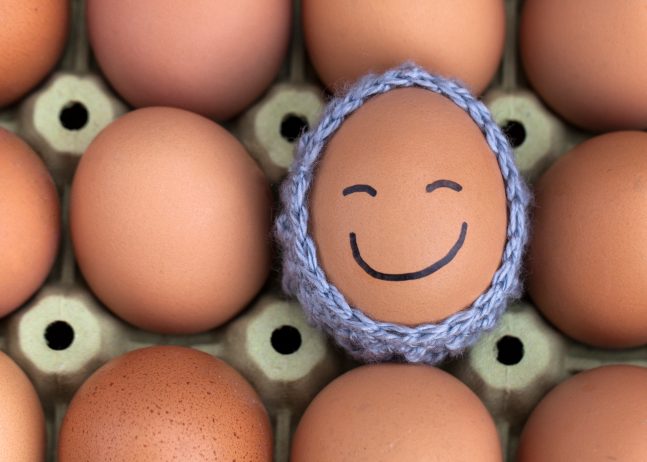 Carton of eggs with one in a knit holder with a smiley face