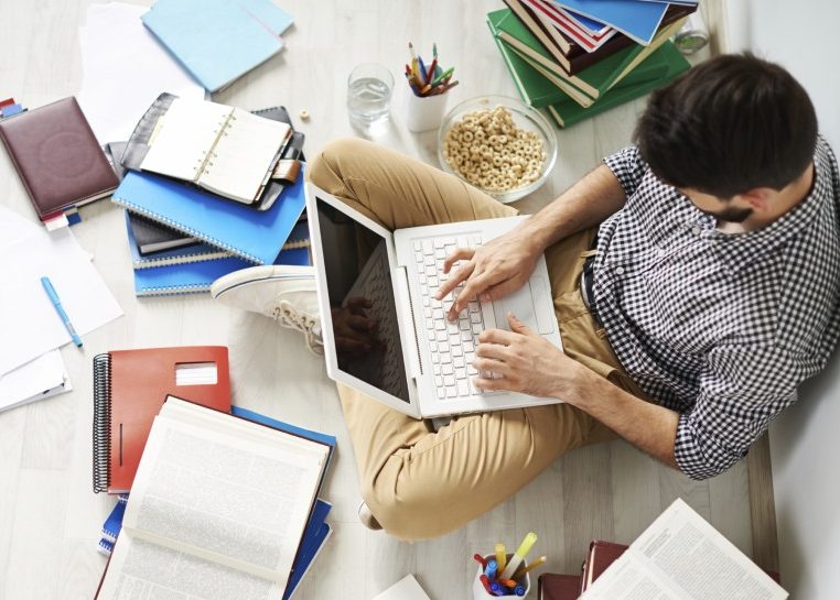 Student sitting on the floor with a laptop in his lap surrounded by notebooks, books, and papers