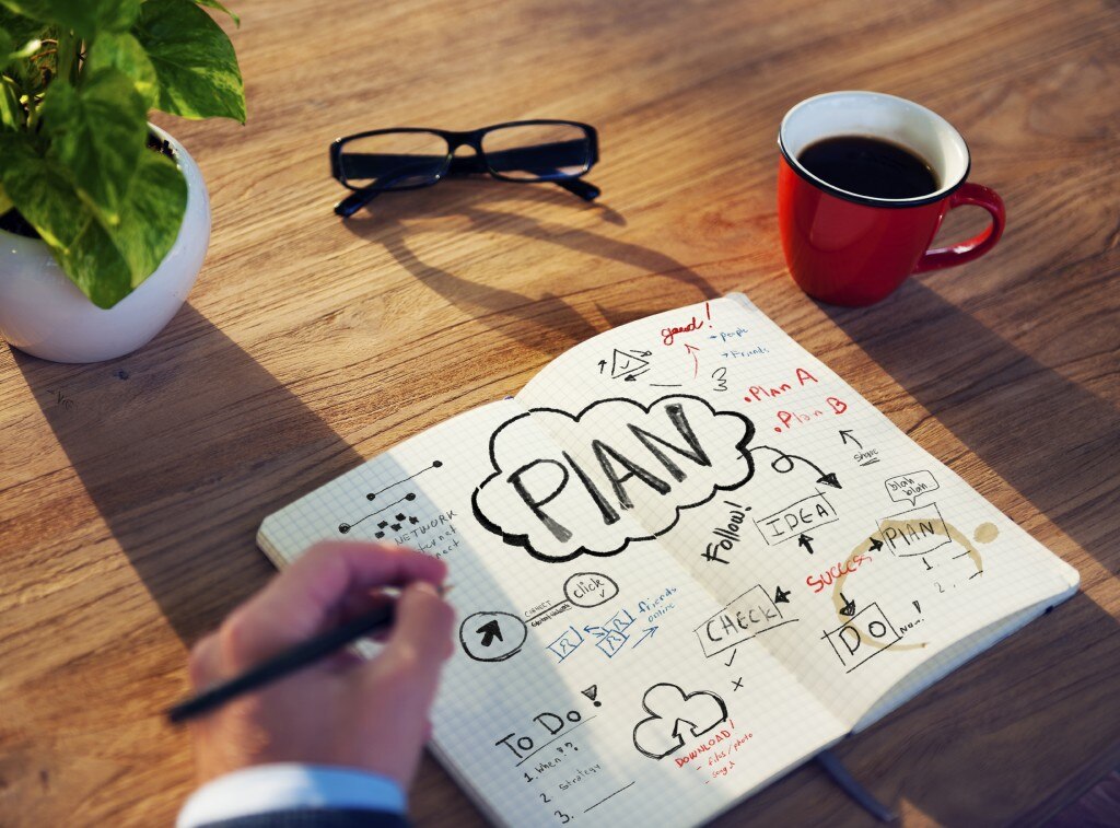 Hand writing in notebook with plans on a desk with a plant, glasses, and cup of coffee