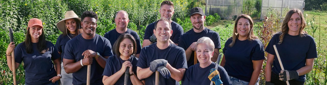 A group of smiling Enterprise Mobility team members holding shovels and rakes standing in a sunny field 